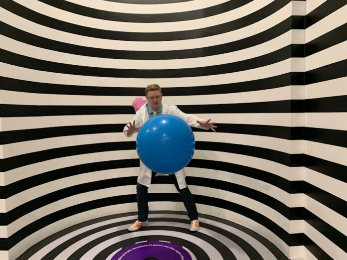 illuseum berlin - A man wearing a lab coat poses with a blue ball in a striped illusion room.